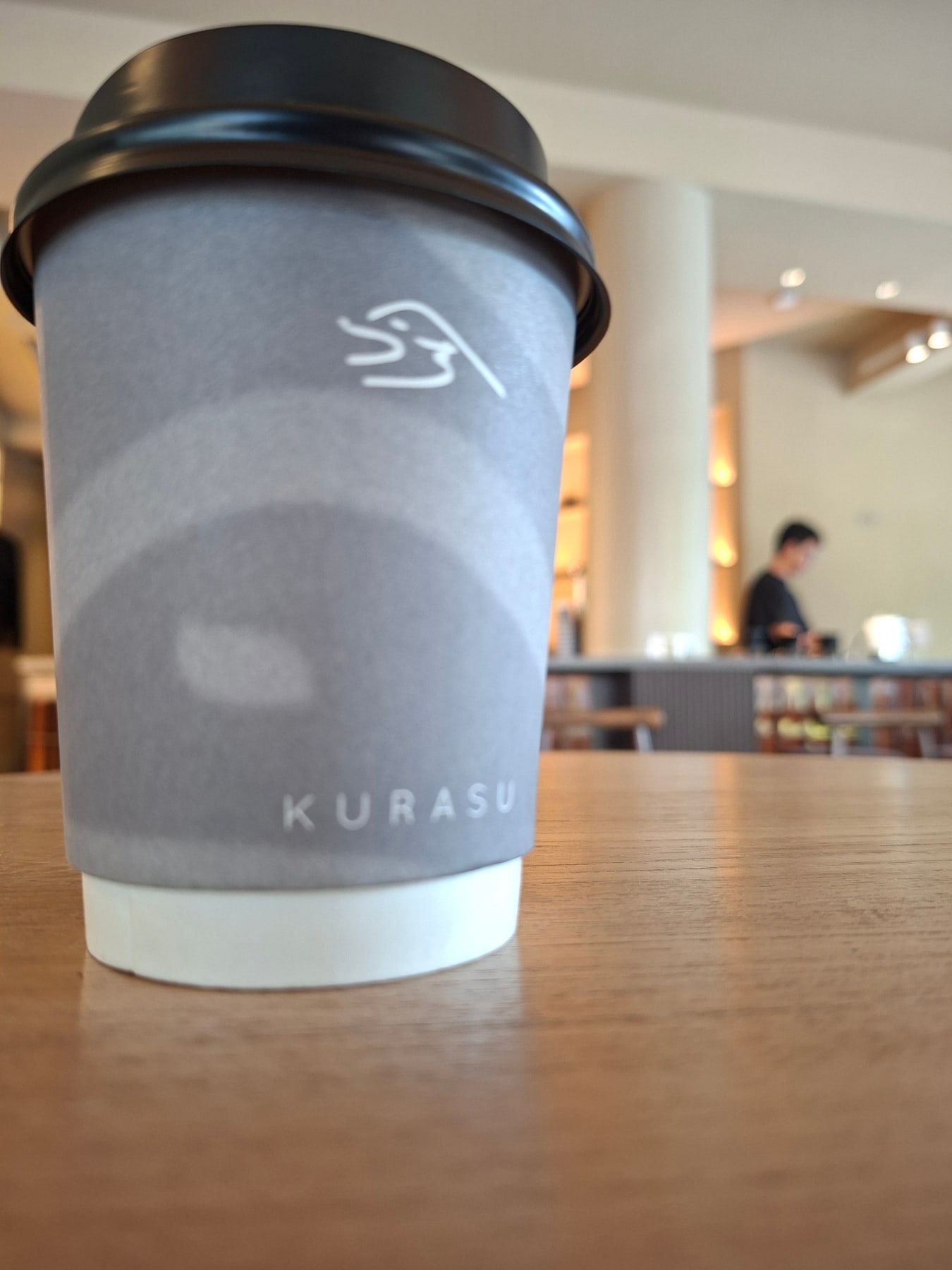 A grey Kurasu takeaway cup on a wooden table inside the Thonglor café